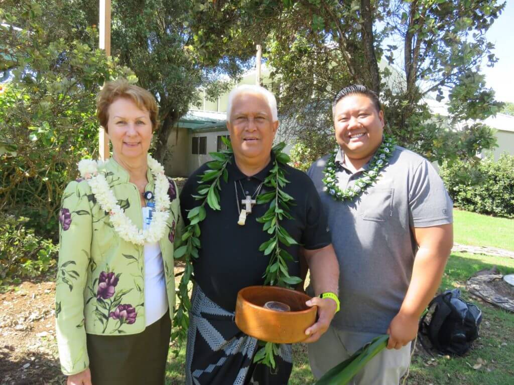 2 Men and 1 woman wearing leis and holding ceremonial items.