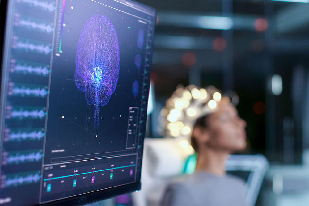 Monitor with high tech brain map with patient in the background with equipment on their head.