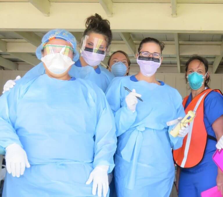 A group of nurses wearing disposable gowns, face masks and shields with one holding thermometer and pen.