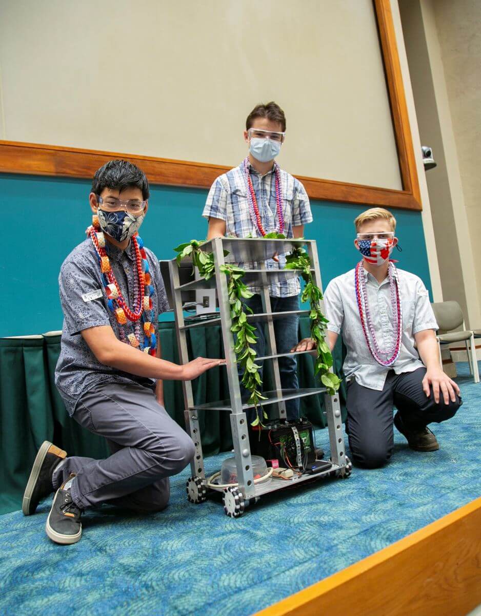 3 students wearing masks and leis standing with a robot that has a lei on.