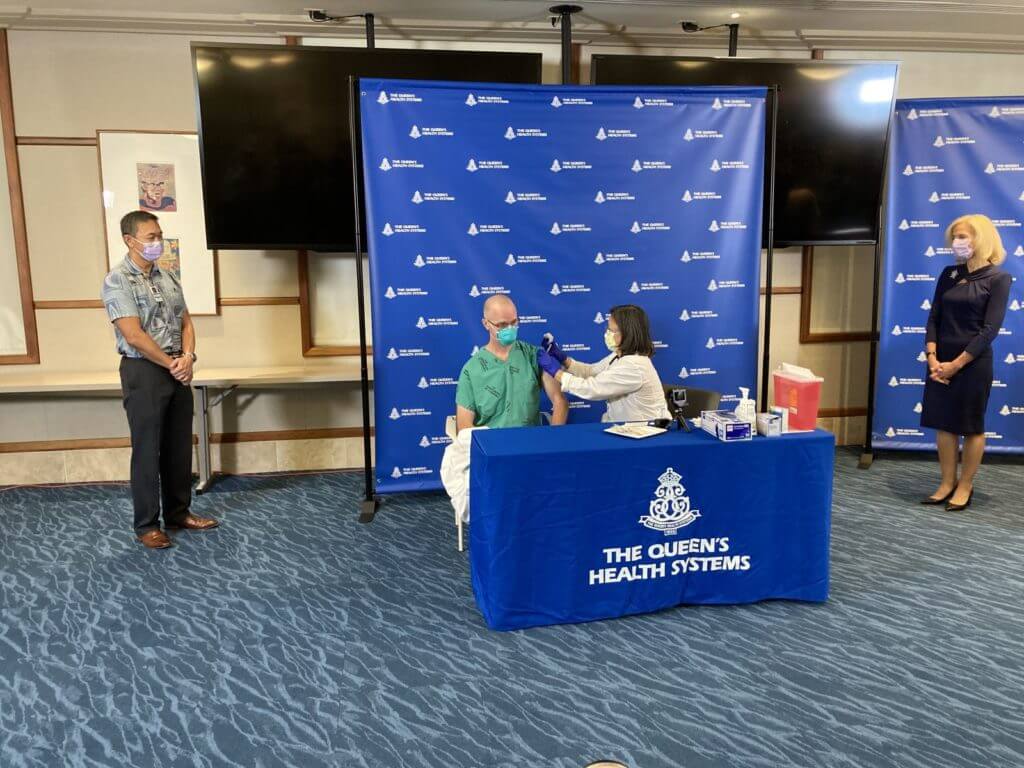 A female doctor administers a vaccine to healthcare worker with QHS leaders watching.