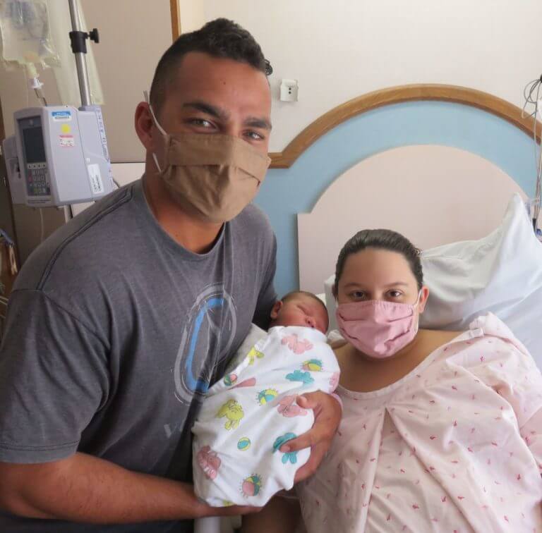 Mother and father with new born baby while wearing masks in a hospital room.