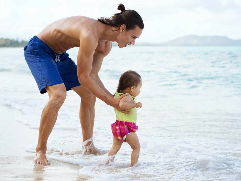 Young man in swim trunks helps toddler stand in the water at the beach.