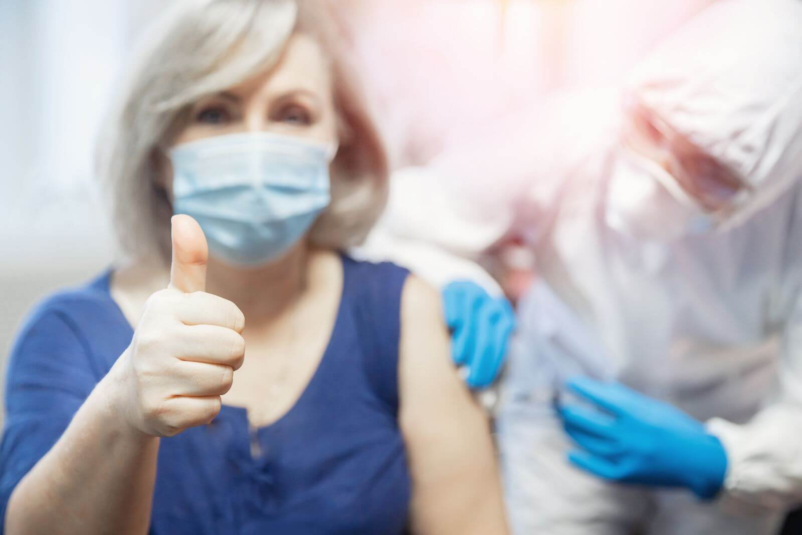 Woman with a facemask receiving a shot while also holding up a thumb.