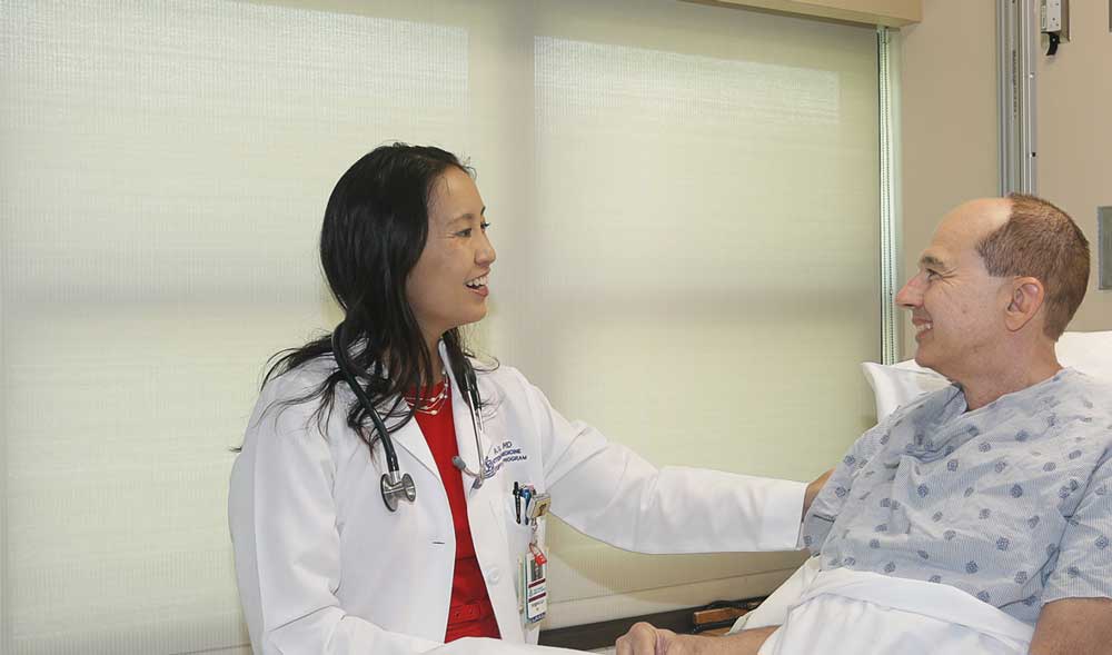 Female Doctor smiles with male patient in hospital bed and gown.
