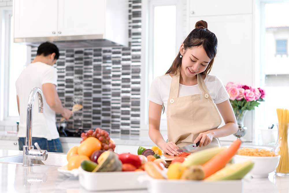 Woman chopping a carrot with a man at cooking something in the background.
